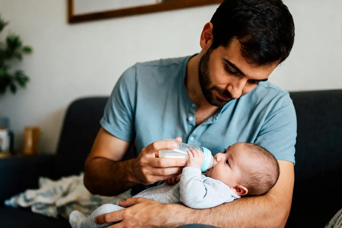 Homme nourrissant un bébé au biberon, assis sur un canapé, dans un intérieur confortable.